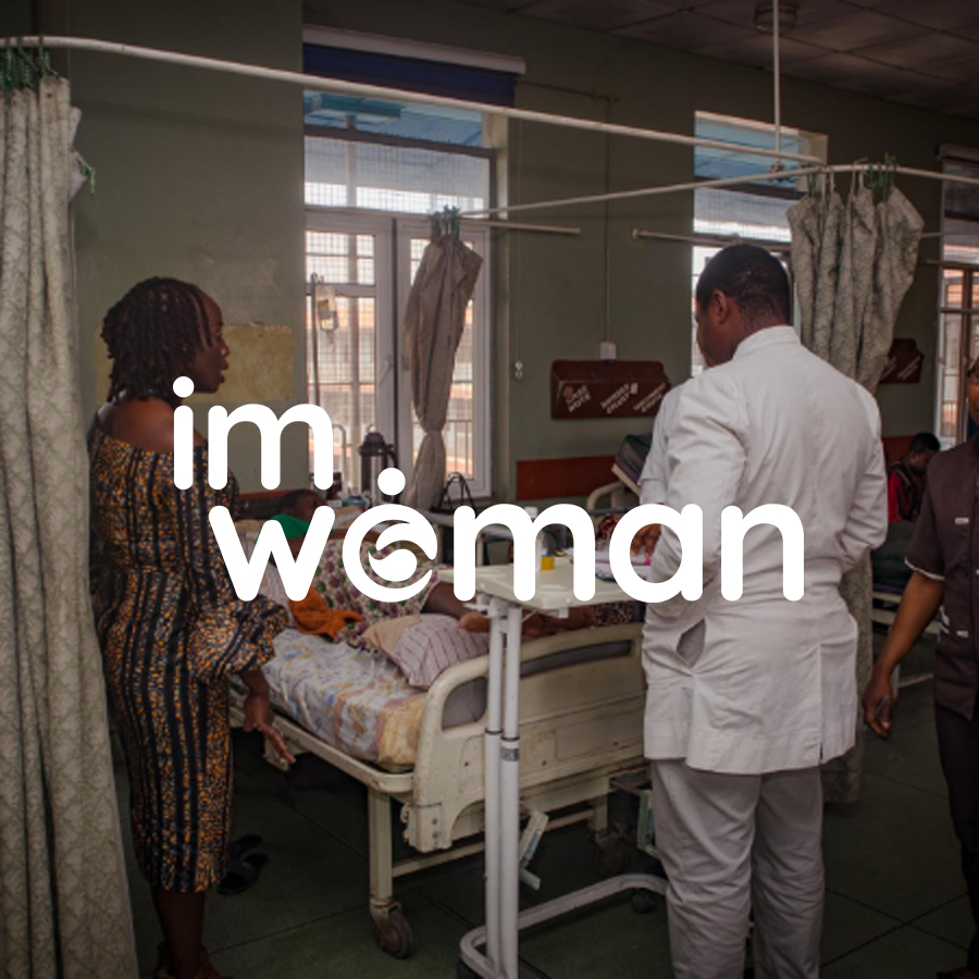A woman in a hospital room stands next to a patient lying in bed, with a healthcare worker present. Text overlay says 'I'm Woman.'