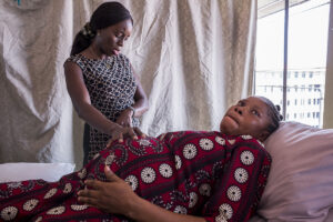 A pregnant woman lies in a hospital bed, a woman stands behind her with one hand placed on the pregnant woman’s stomach while looking towards her watch.