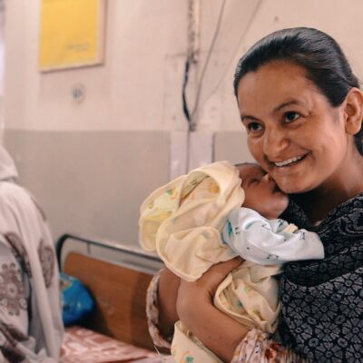 A mother in a hospital holding her newborn baby while smiling, with other patients visible in the background