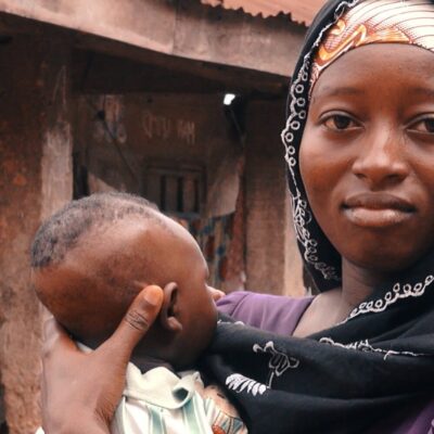 A woman in a headscarf holding a baby outside in a rural setting, looking directly at the camera