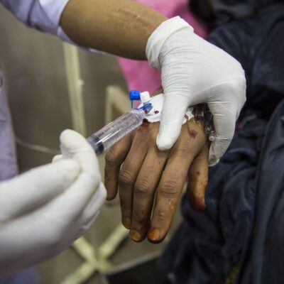 A close-up of a healthcare worker wearing gloves, administering an intravenous (IV) injection to a patient’s hand