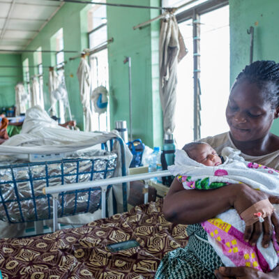 A mother holding her baby in a hospital setting with multiple beds.